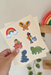 Colorful stickers featuring a rainbow, butterfly, child in a rocket, and dinosaur held by a hand on a light background.